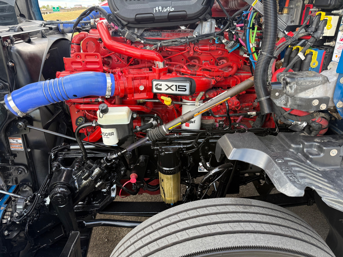 Kenworth T680 engine bay with red engine block, X15 badge, blue intercooler hose, white coolant reservoir, and various black hoses and wiring in view.