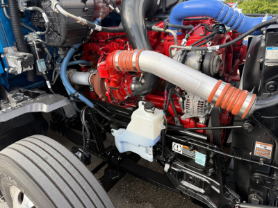 Kenworth T680 engine bay showing red engine block, large intake turbo piping, blue coolant hose, white reservoir, and various hoses and components.