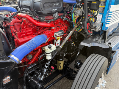 Kenworth T680 engine bay showing red engine with blue silicone intake hose and EXIS badge, various hoses, wiring harnesses, and engine components.