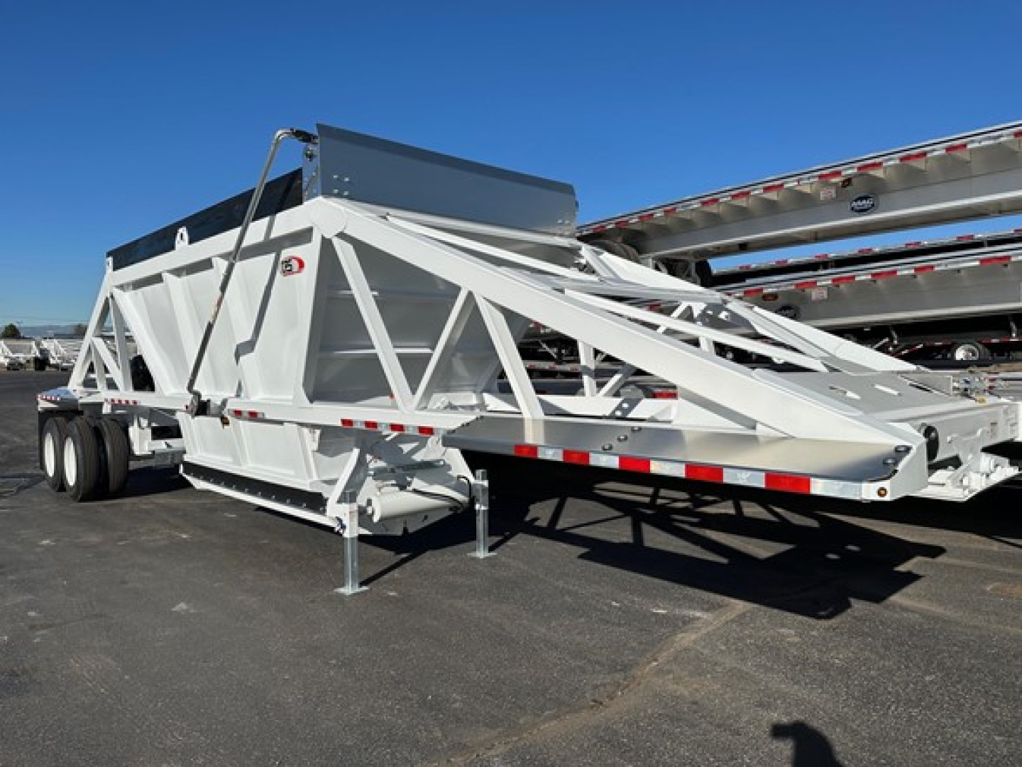 Bottom-dump trailer on a concrete lot, white framework with triangular truss structure and a top hopper, cabless front appears to be folded, with a black tank or chute along the upper rear.