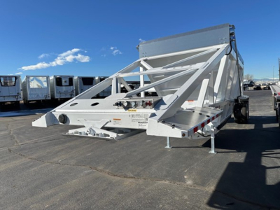 Bottom-dump trailer in white with a structural truss frame, side supports, and a cab-over style underside mechanism. Clear sky and other trailers in background.