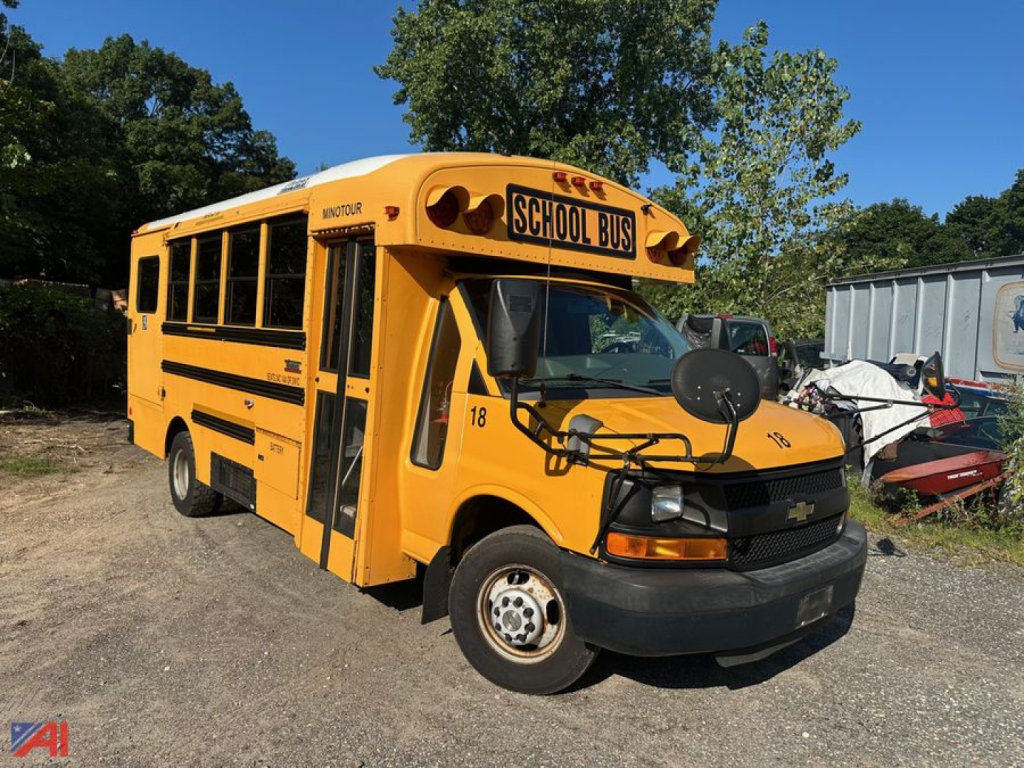 Yellow school bus body with wheelchair lift; 'SCHOOL BUS' sign on top; Chevrolet front; number 18 on door; black bumper; side doors and windows; outdoor lot with trees.