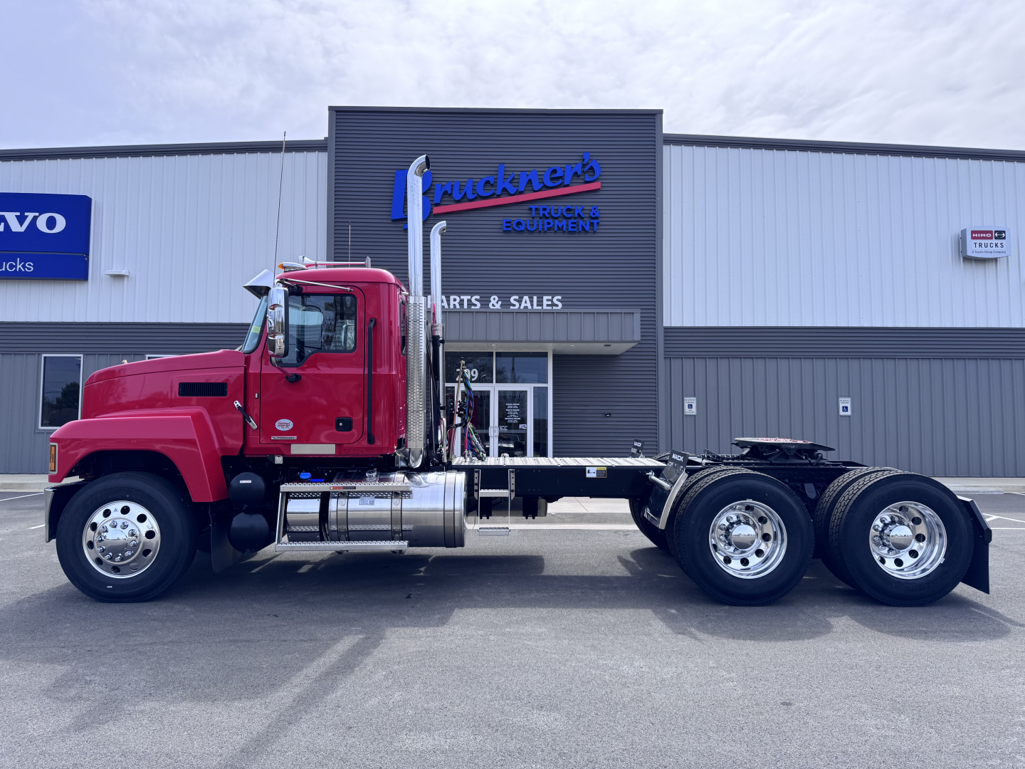Truck chassis visible from left side with bright red cab, long wheelbase, dual rear axles, and fuel tanks; dealership building in background with 'Truckers' logo.