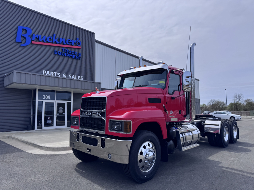 Mack PI64T conventional daycab truck front-left view in dealership lot with chrome bumper, dual rear axles, red cab, and Bruckner's Trucks sign in background.
