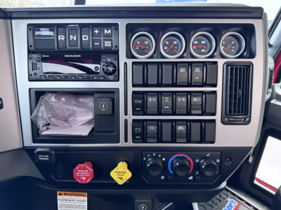 Truck dash panel showing gauges, switches, and radio in Mack PI64T cab; multiple round gauges, control knobs, ignition area, and warning buttons.