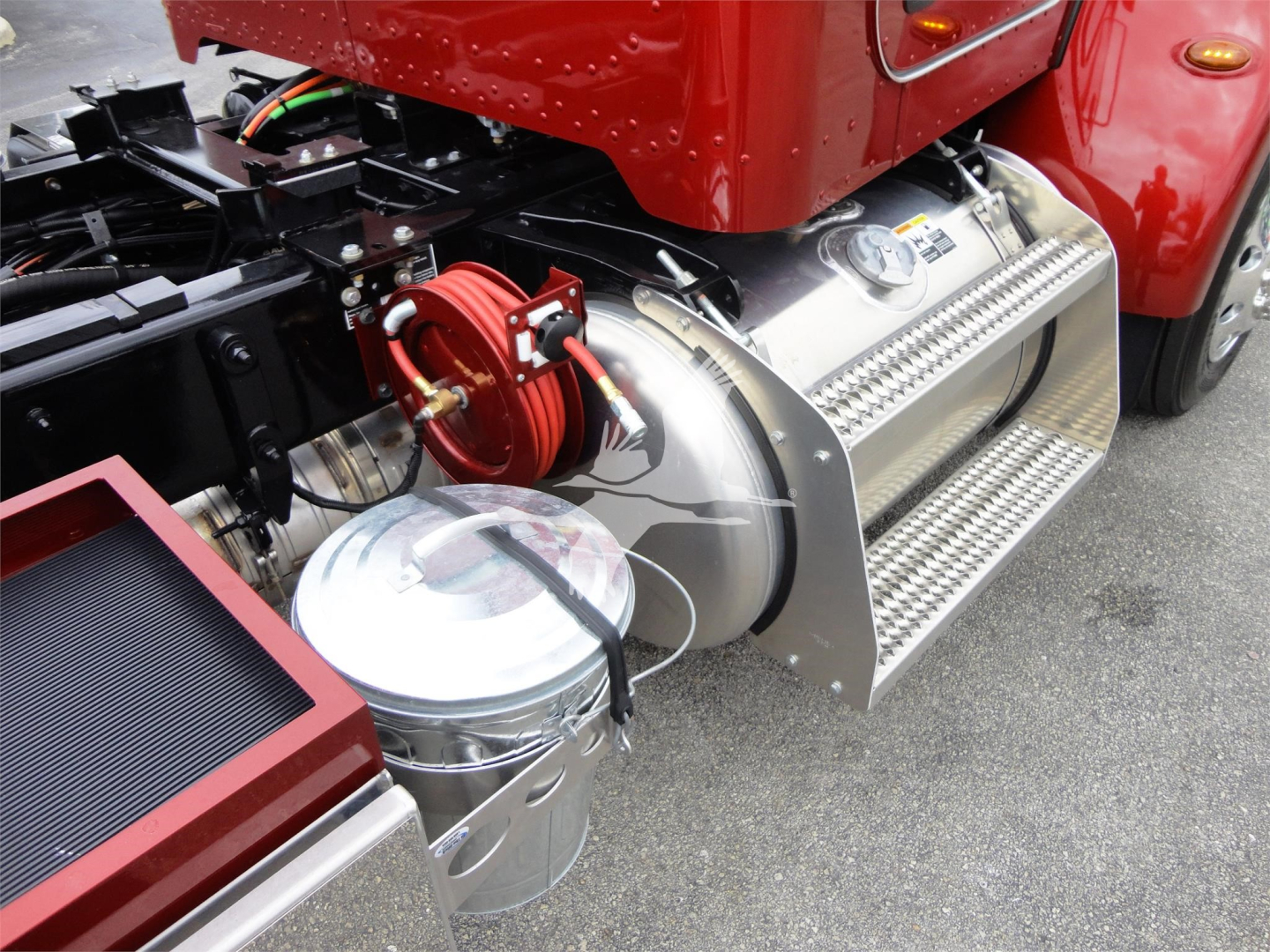 Truck side view showing chrome fuel tank and aluminum step aboard on a red Peterbilt 337 with a coiled hose reel and cylindrical canister nearby.