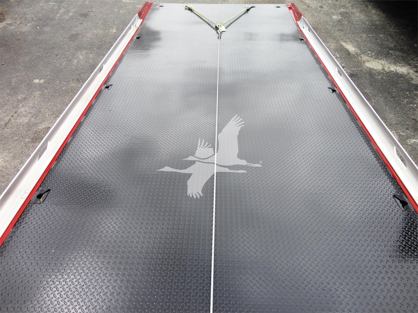Flatbed/rollback truck bed with diamond plate surface and a bird logo in center, silver rails along edges, and a tie-down strap at the front.