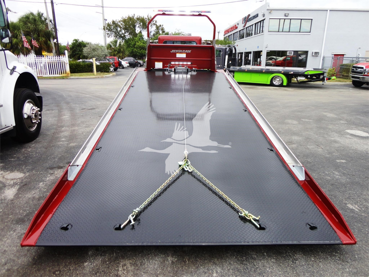 Peterbilt 337 rollback tow truck bed with black embossed deck and red side rails, center winch anchor chains securing the surface, dealership lot in background.