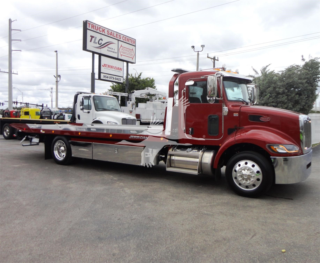 Peterbilt 337 rollback tow truck in red with chrome accents, long heavy-duty flatbed; dealership sign in background; visible text includes TRUCK SALES CENTER on sign and Peterbilt badge on truck.