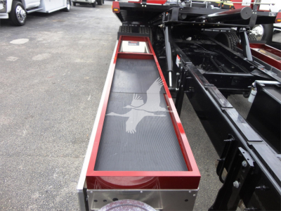 Truck chassis with a red-bordered, black ribbed belt-like platform (rollback bed) extending toward camera; Peterbilt 337 equipment visible; metal frame and hydraulic components in background.