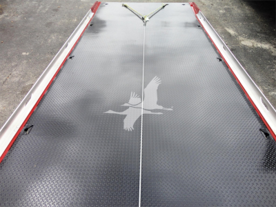 Flatbed/rollback truck bed with diamond plate surface and a bird logo in center, silver rails along edges, and a tie-down strap at the front.