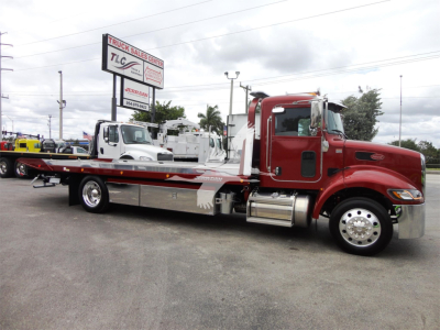 Peterbilt 337 rollback tow truck side view with red cab and flatbed, chrome exhaust stack, and wheel covers; dealership lot in background.