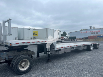 Trailer image showing a long flatbed drop deck trailer with railing and hydraulic landing gear. Visible text includes 'Drop Deck' and 'Reitnouer' branding on the equipment, with a cloudy sky in background.