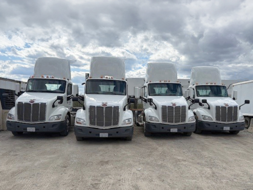 Three Peterbilt 579 trucks lined up side-by-side with raised sleeper cabs, front view, in a lot under cloudy sky.