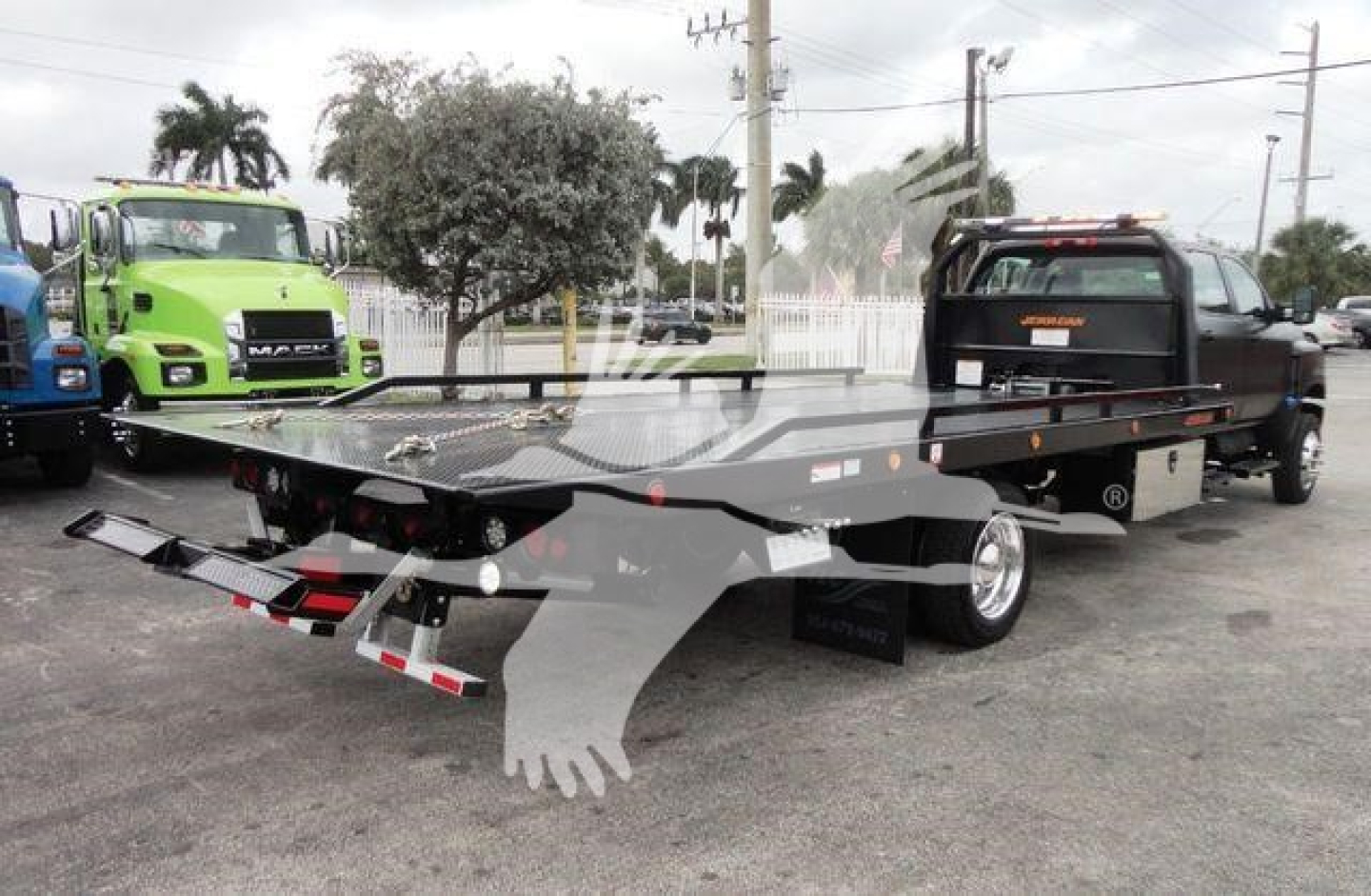 Flatbed rollback tow truck with black bed and rear ramps, black cab, and green truck in background. Visible text includes stock number J98978 and category/type signage in listing context.
