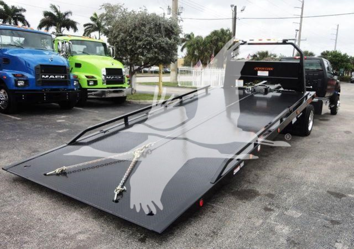 Heavy-duty rollback tow truck with a long flatbed in the foreground, black metal surface, two hydraulic ramps, and a pickup bed with a rear-mounted ramp; multiple colorful trucks in background.