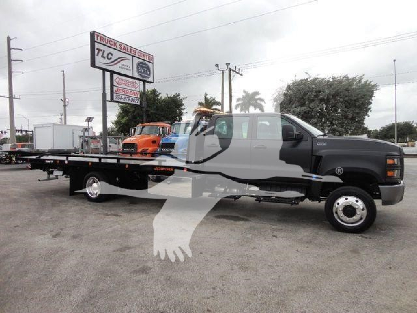 Flatbed tow truck with logo overlay showing a long black cab and silver wheels, rollback truck in a dealership lot.