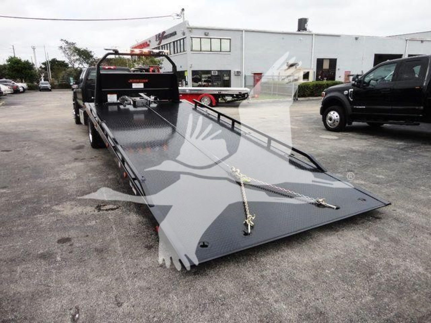 Flatbed tow truck trailer with a long gray ramp-style bed, front cab, and chains securing the deck; parking lot with other trucks in background.