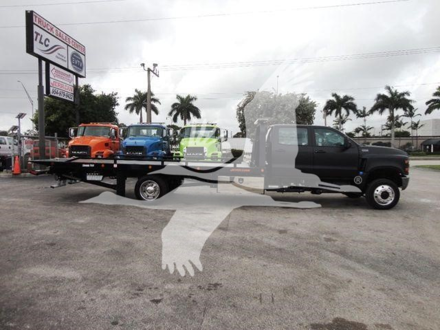 Flatbed tow truck with several colorful trucks on the flatbed in a dealership lot. TLC Truck Sales sign visible in background. Overlaid watermark of two hands shaking.