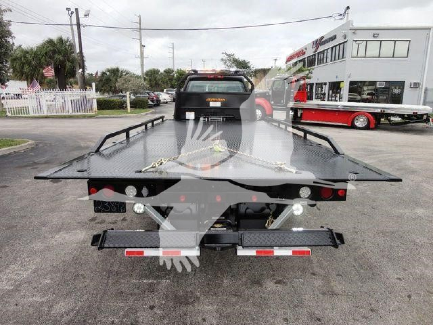Flatbed tow truck bed with ramps and safety chains, seen from rear, in a dealership lot.