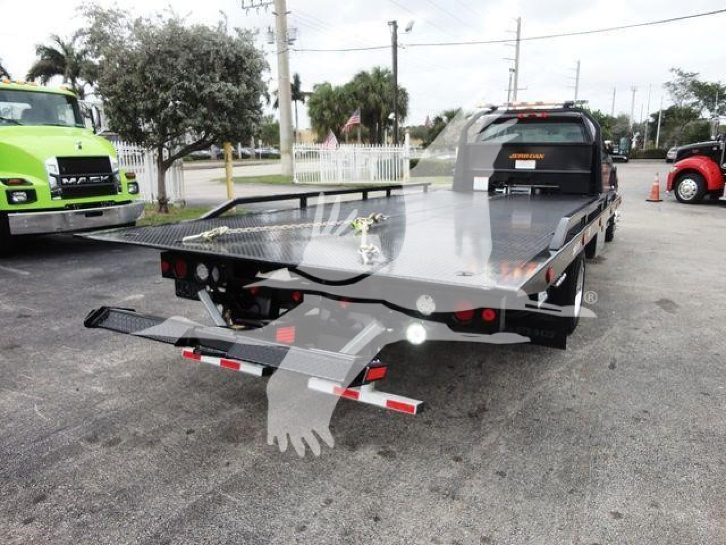 Flatbed tow truck with rear-view showing metal deck and ramp, International trailer, in-service scene with other trucks in background.