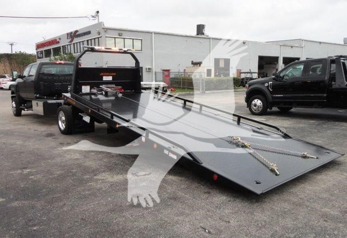 Flatbed rollback tow truck with a long loading ramp, parked in lot; background shows a service building and other trucks.