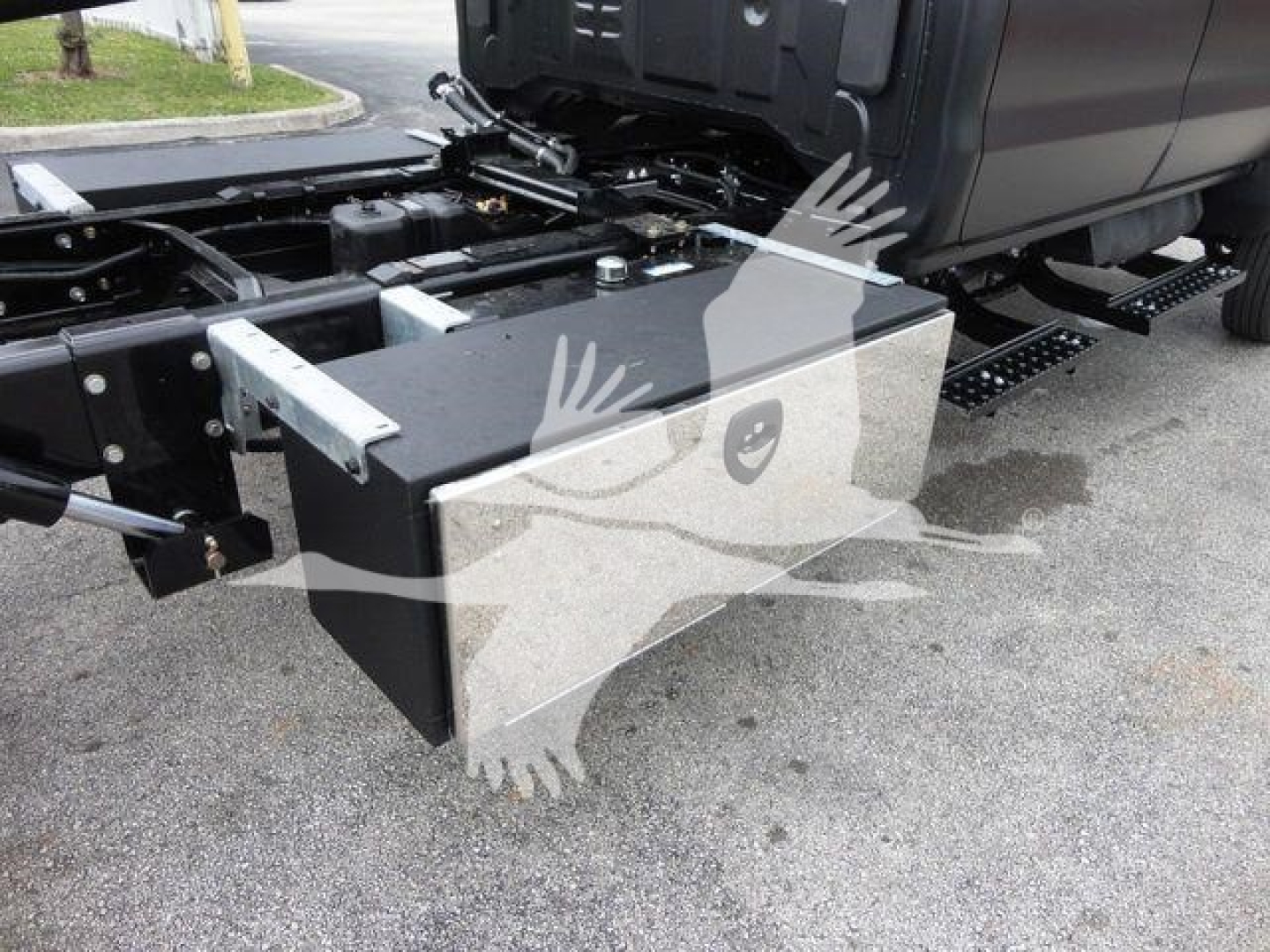 Truck chassis side view showing a large rectangular black toolbox mounted near the front of the cab with metal brackets and a hydraulic line nearby.