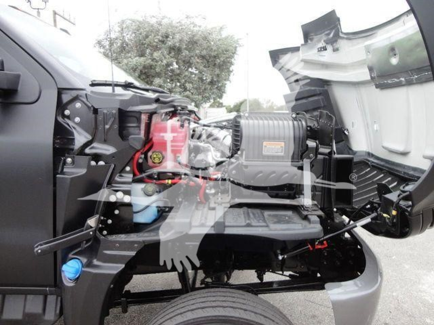 Truck cab interior showing engine area with exposed components and a red reservoir, all under a raised hood.