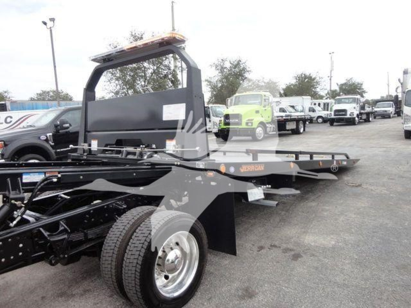 Truck rollback bed with frame and wheels visible; flatbed with hydraulic components and upright railing; background shows dealership lot with multiple trucks.