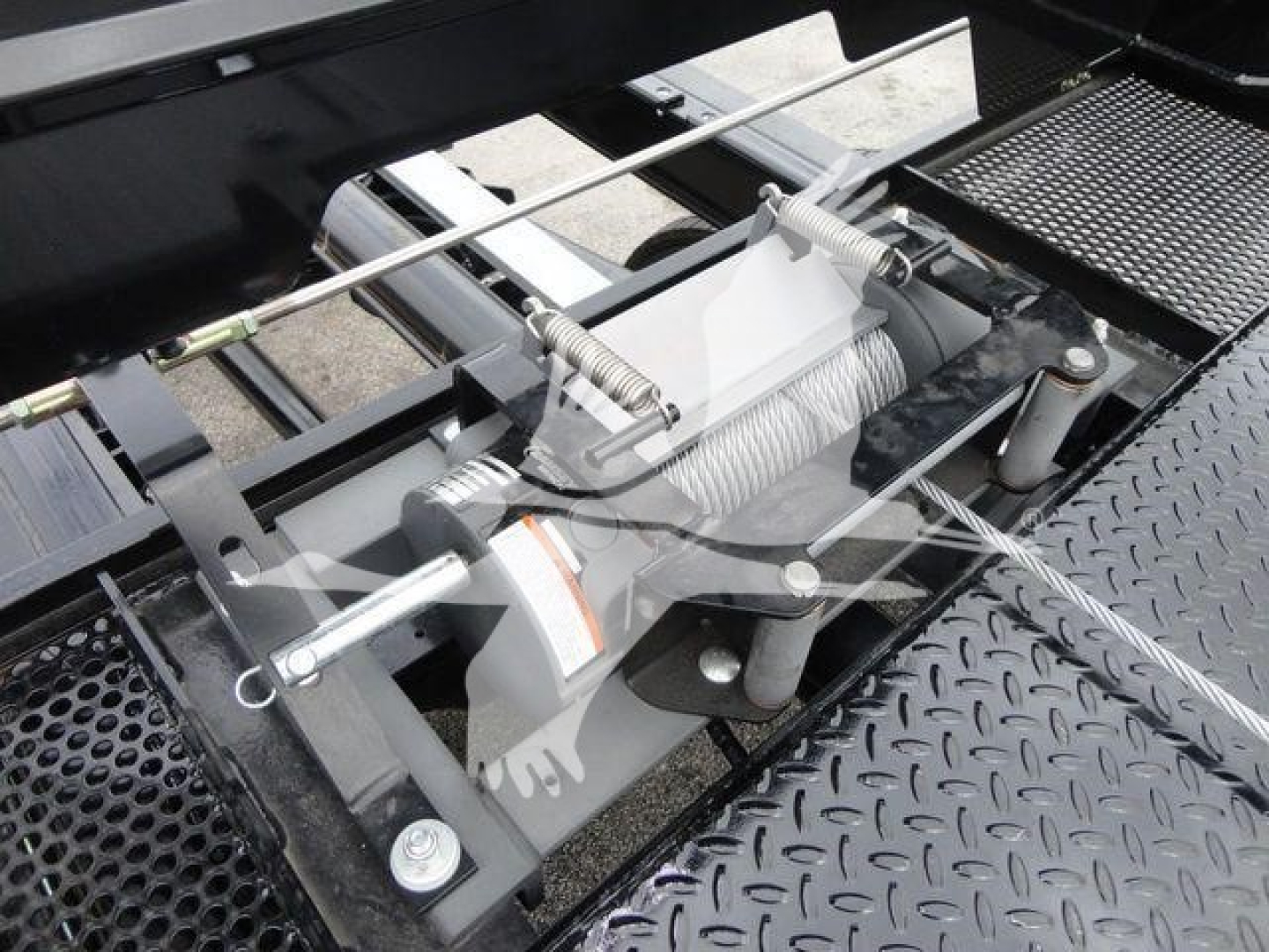 Trailer winch and mechanism assembly mounted on a diamond plate platform; visible components include a spring-loaded cable drum, steel pins, and a hydraulic-like bracket with bolts.