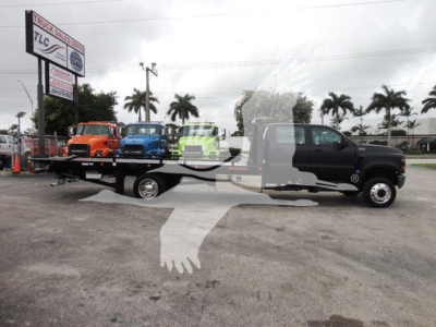 Flatbed tow truck with several colorful trucks on the flatbed in a dealership lot. TLC Truck Sales sign visible in background. Overlaid watermark of two hands shaking.