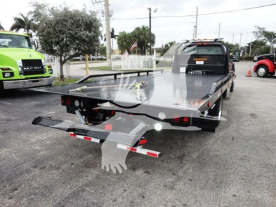 Flatbed tow truck with rear-view showing metal deck and ramp, International trailer, in-service scene with other trucks in background.