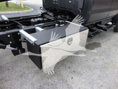 Truck chassis side view showing a large rectangular black toolbox mounted near the front of the cab with metal brackets and a hydraulic line nearby.