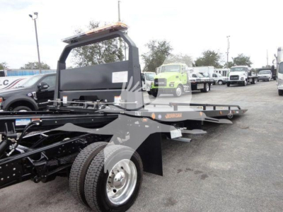 Truck rollback bed with frame and wheels visible; flatbed with hydraulic components and upright railing; background shows dealership lot with multiple trucks.