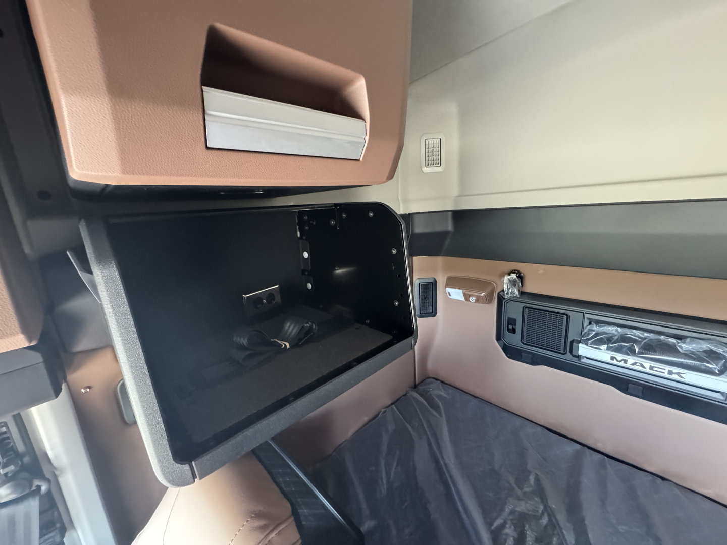 Truck cab interior showing open overhead storage compartment and dashboard with Mack branding and tan/brown trim.