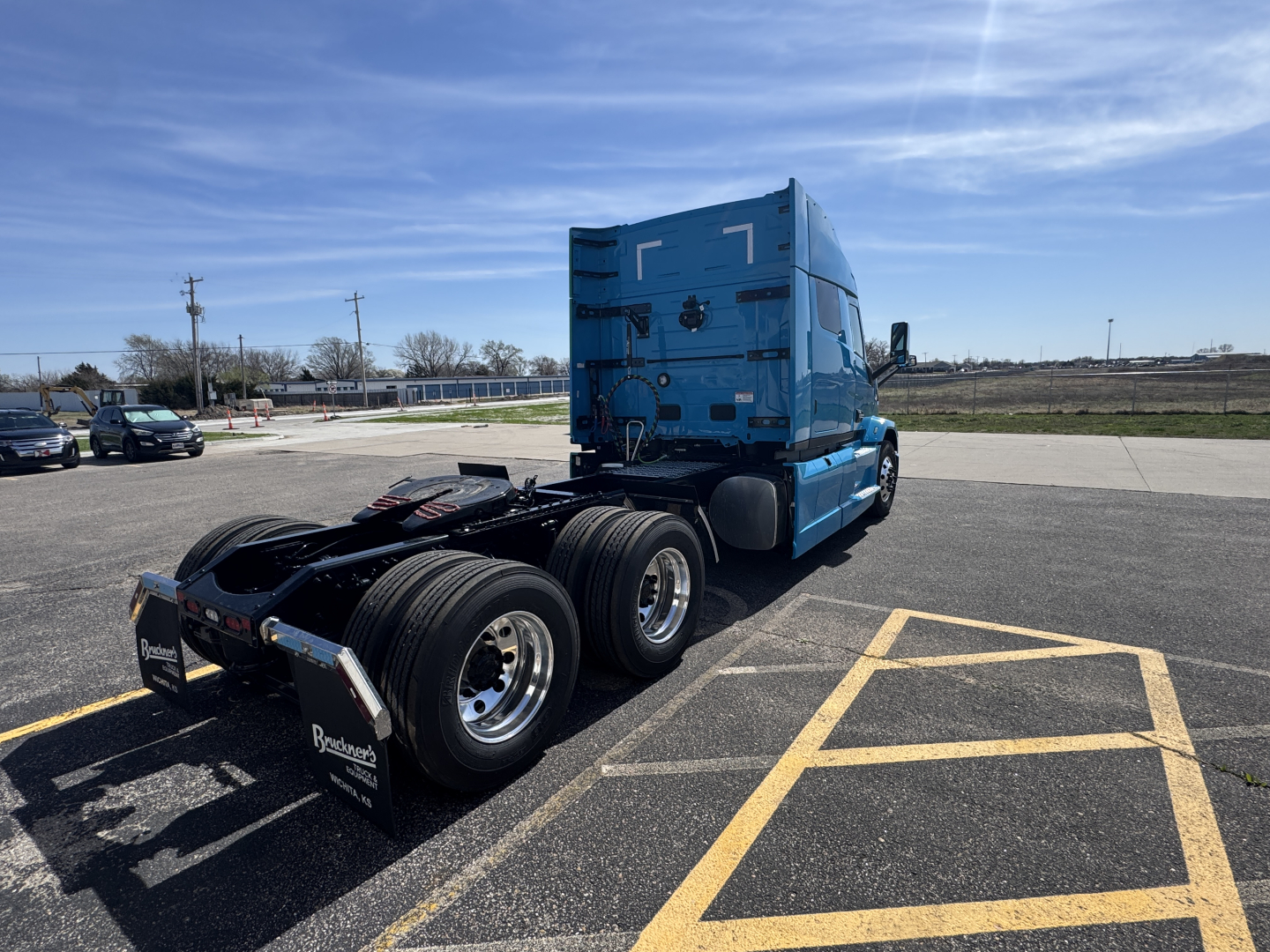 Truck tractor with blue cab and dual rear axles in a parking lot, rear view showing fifth wheel area and dual rear tires.