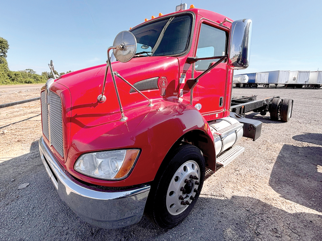Kenworth T370 cab and chassis, red exterior, front view showing side mirror, headlight, and chrome bumper; front wheel and dual rear wheels visible; daylight outdoor lot.