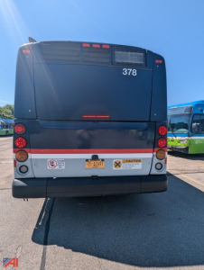 Back view of a 2013 New Flyer XD60 transit bus showing rear bumper, taillights, and yellow warning labels.