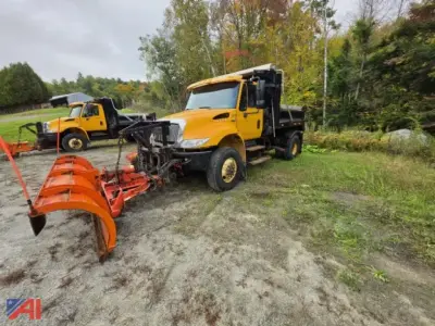 Dump truck with plow and wing attachment in a grassy lot; yellow International 4300 with black plow blade and orange wing plow in foreground.