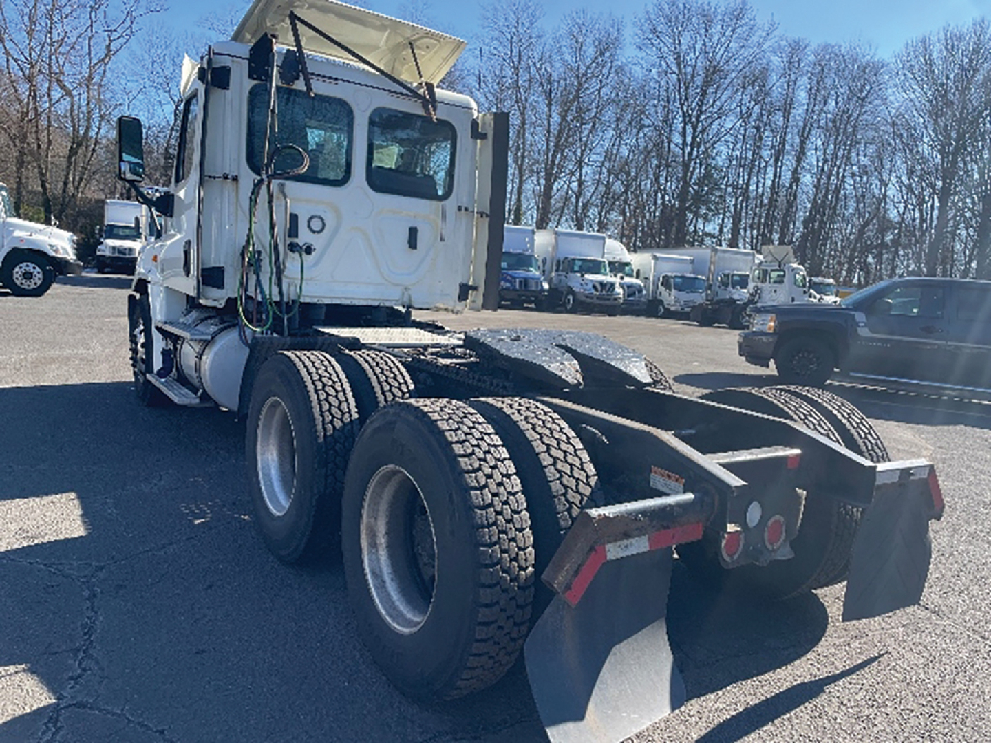 Freightliner Cascadia125 day cab rear/side view showing dual rear axles and fifth wheel with mud flaps. White cab with overhead exhaust, exposed hoses, and parked trucks in lot.