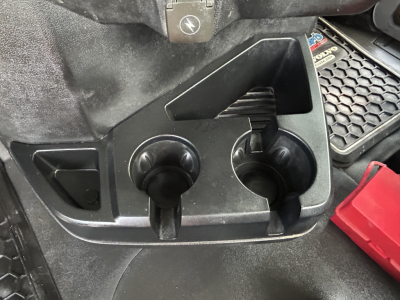 Two circular cup holders and a storage area on the Mack GR64B cab interior noting a red tool or component on the right; visible plastic panels and textured floor mat