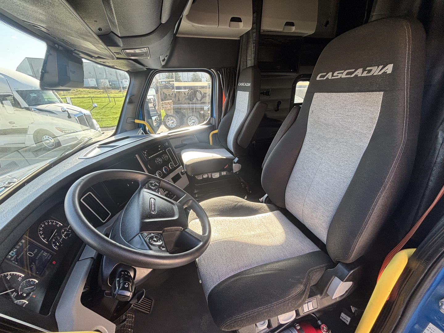 Truck cab interior showing driver seat with 'CASCADIA' upholstery, steering wheel, dashboard, and passenger seat; cab window and overhead storage visible.