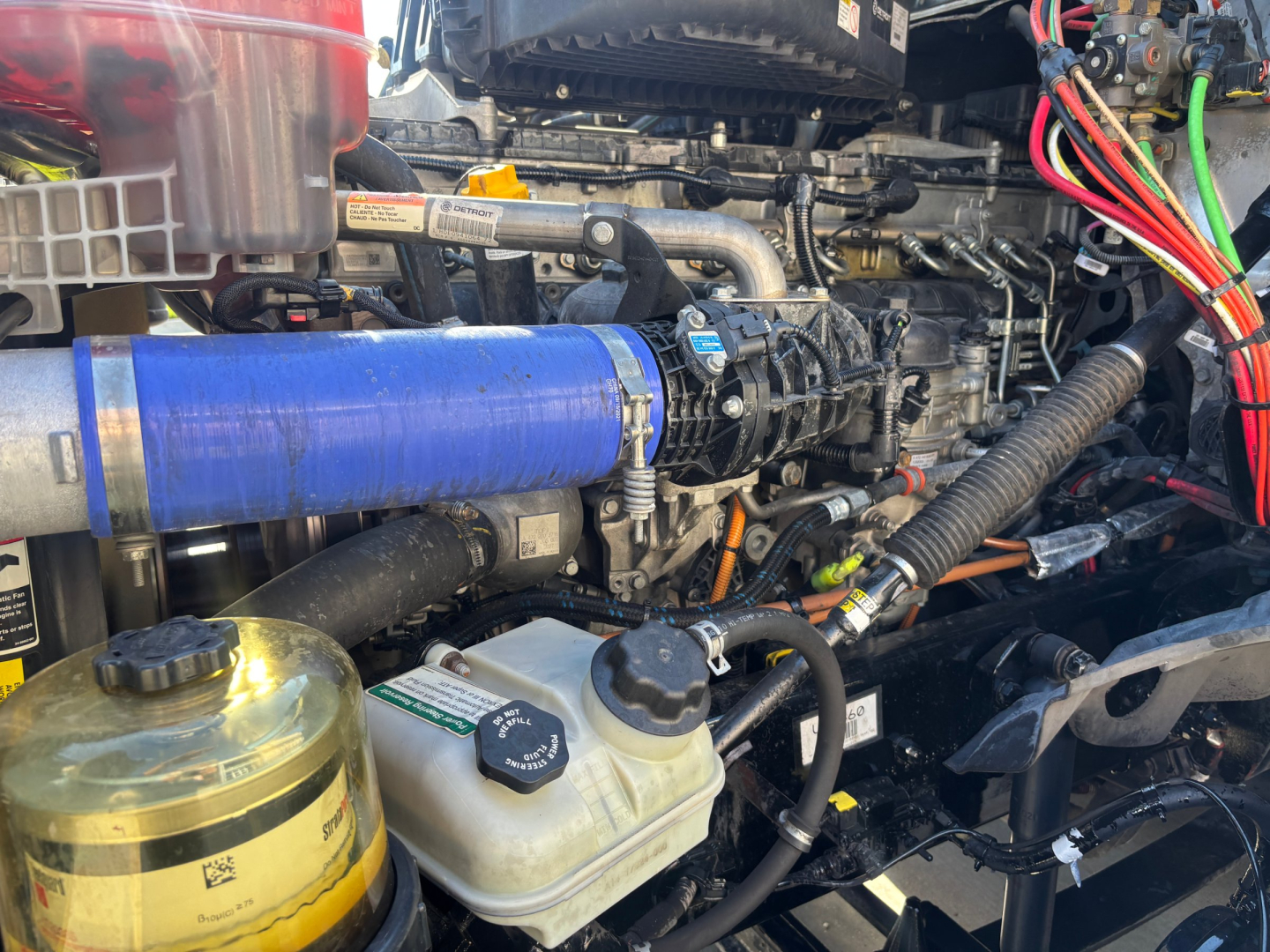 Truck engine bay with blue silicone hose, coolant reservoir, and various hoses and wiring in a Freightliner PT126064ST engine compartment.