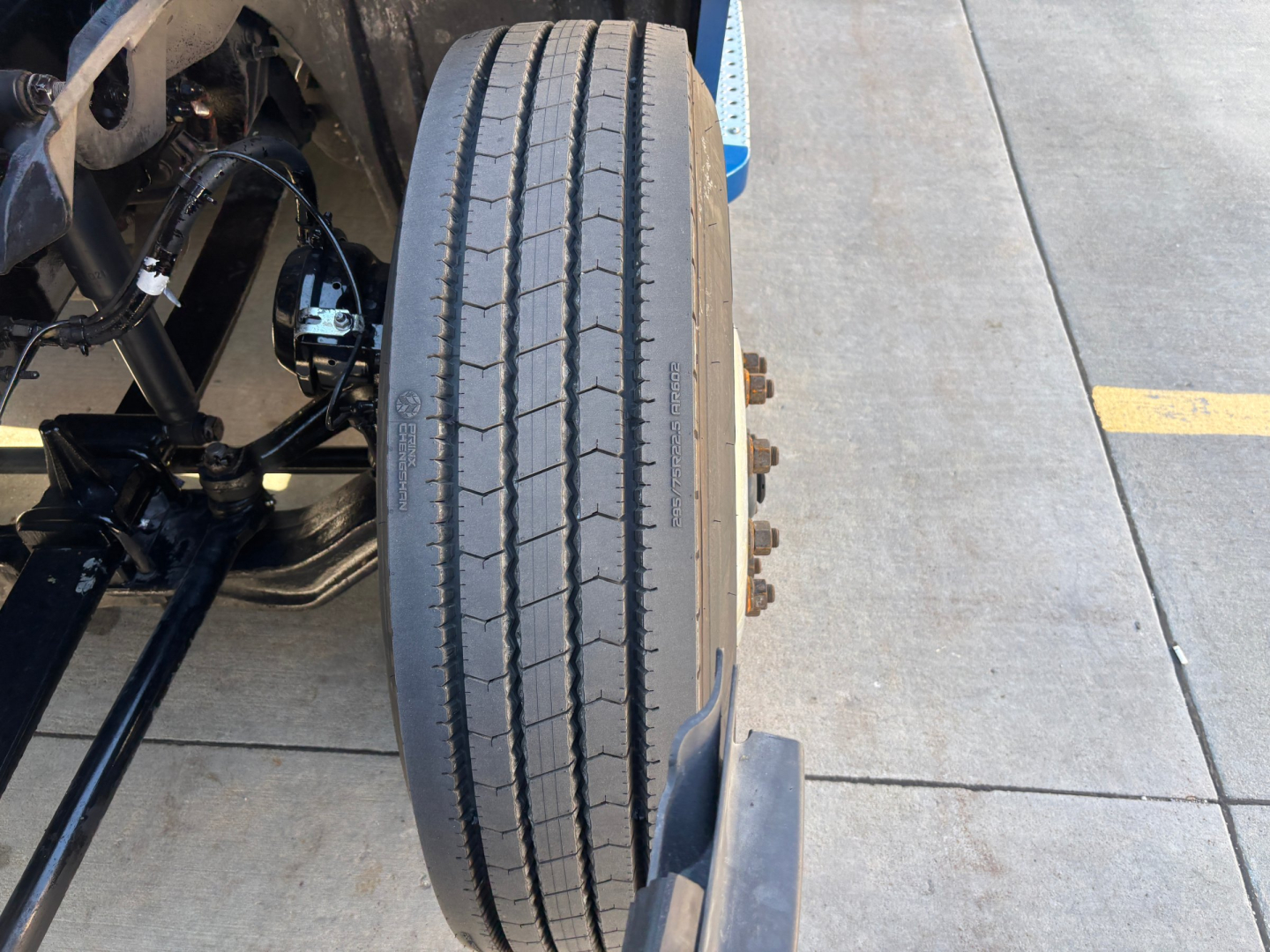 Truck front axle with tire and suspension components visible; tread pattern on steering axle tire; part of wheel hub and brake assembly.