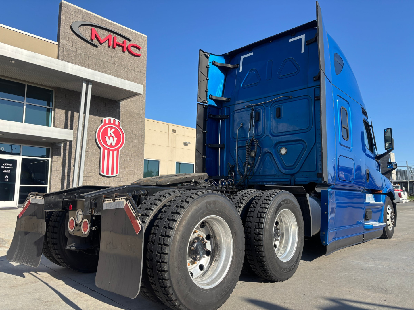 Freightliner PT126064ST tractor rear rear-axle view showing dual rear wheels and mudflaps; blue sleeper cab with stacked exhaust and trailer hitch, dealership building in background.