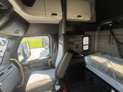Cab area of a Freightliner truck sleeper with overhead storage and bunk, captain seat, and window view; inside shows bed area with mattress and partitions.
