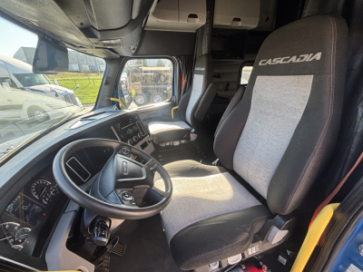 Truck cab interior showing driver seat with 'CASCADIA' upholstery, steering wheel, dashboard, and passenger seat; cab window and overhead storage visible.