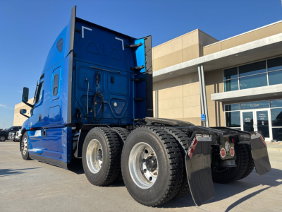 Multiple-axle Freightliner tractor in blue, rear view showing dual rear tandem wheels and mud flaps, cab with sleeper, dealership building in background.