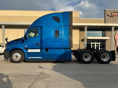 Truck side view showing a blue Freightliner PT126064ST conventional sleeper tractor with dual rear axles and black accents parked in front of a building.