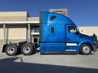 Truck side view of a blue Freightliner PT126064ST conventional sleeper with three-axle rear, parked outside a building.